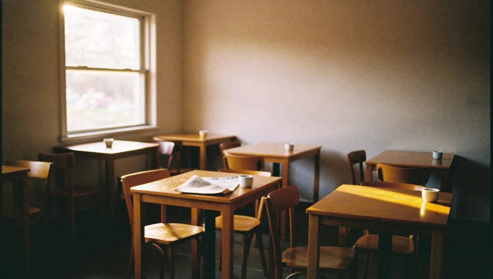 A wide view of a small, plainly furnished cafe room with wooden tables and daylight from the side window